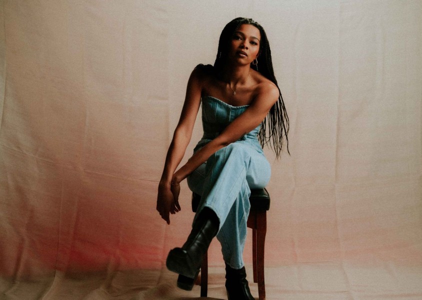 A woman with ling braided hair in a denim jumpsuit sat on a stool x